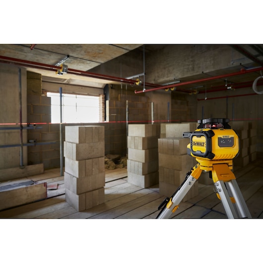 A DEWALT laser level device mounted on a tripod is positioned in a partially constructed room with concrete block walls and exposed pipes on the ceiling. The laser projects a horizontal red line across the room, aiding in alignment during construction.