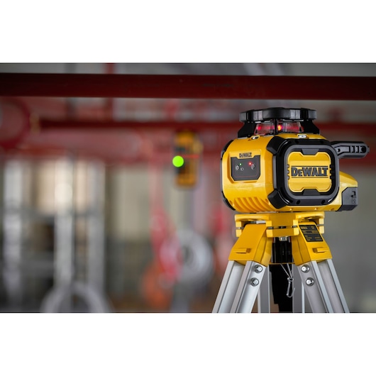A DEWALT DCLE05211R yellow laser level mounted on a tripod, positioned in an industrial setting with red pipes and blurred background.