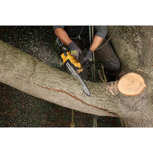 A person is using a DEWALT cordless chainsaw to cut a thick tree branch. Wood chips are flying, and the person is wearing protective gloves and gear.