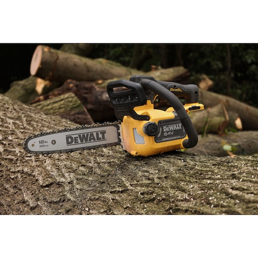 A DEWALT 54V cordless chainsaw rests on a large tree trunk outdoors, surrounded by cut logs. The chainsaw features a 12-inch (30 cm) blade and is yellow and black in color.
