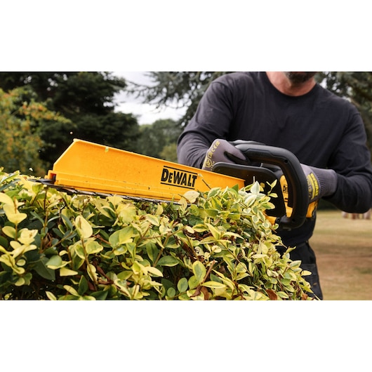A person wearing gloves is using a DEWALT cordless hedge trimmer to trim green bushes outdoors.