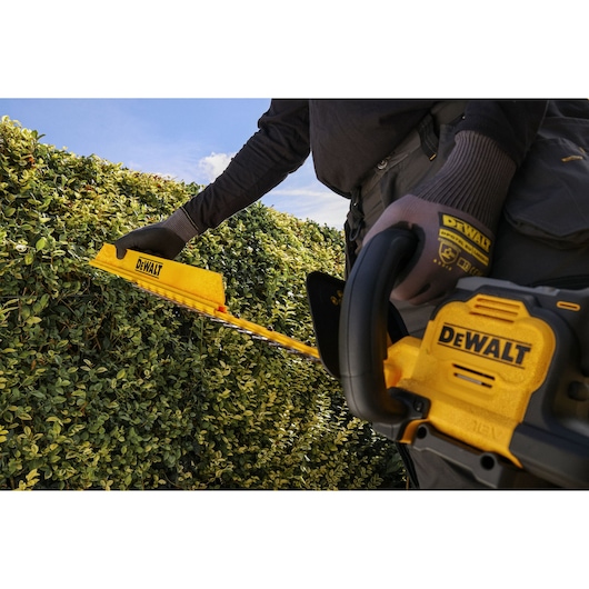 A person using a DEWALT cordless hedge trimmer to trim a green hedge outdoors under a clear sky.