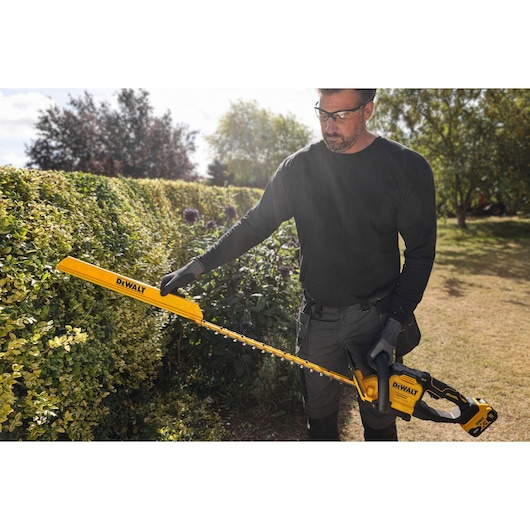 A person using a DEWALT cordless hedge trimmer to trim bushes outdoors in a garden. The hedge trimmer is yellow and black and the person is wearing gloves and workwear.