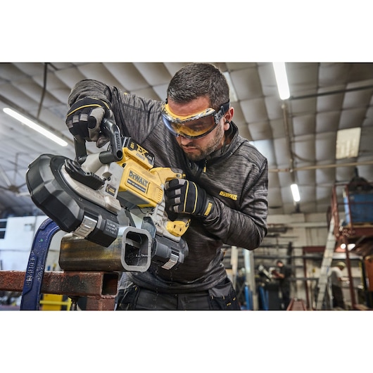 Person using a DEWALT DCS374N portable band saw to cut a metal pipe in an industrial workshop. The individual is wearing protective gloves and a dark jacket with DEWALT branding.