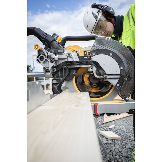 The DEWALT 18V XR 305mm Slide Mitre Saw being used by worker with safety goggles, hat and ear defenders to cut through timber beam outdoors