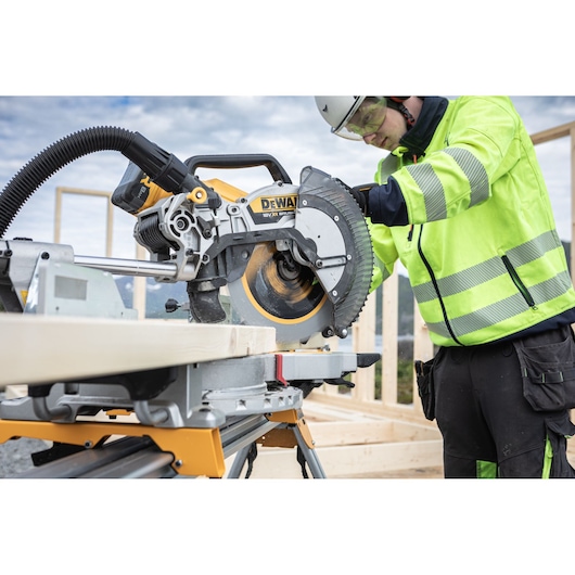 The DEWALT 18V XR 305mm Slide Mitre Saw being used by worker with safety goggles, hat and ear defenders to cut through timber beam outdoors