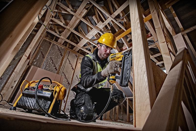Worker uses DEWALT nail gun with compressor in a wooden construction site, wearing safety gear.