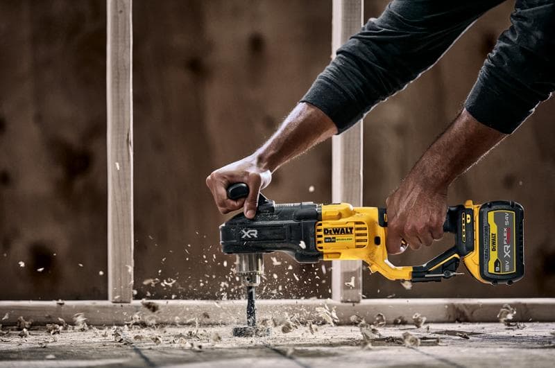 A person using a DEWALT DCD444N cordless power drill with a side handle to drill through wood, creating wood shavings and dust. Construction framing is visible in the background.