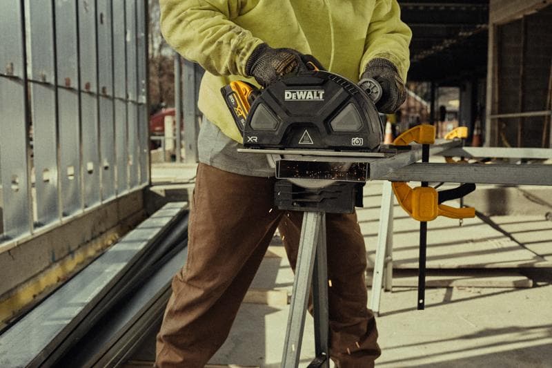 A person wearing work gloves and a yellow sweatshirt is using a DEWALT cordless circular saw to cut metal at a construction site. Sparks are visible as the saw slices through the metal, and the person is operating the saw on a workbench outdoors.